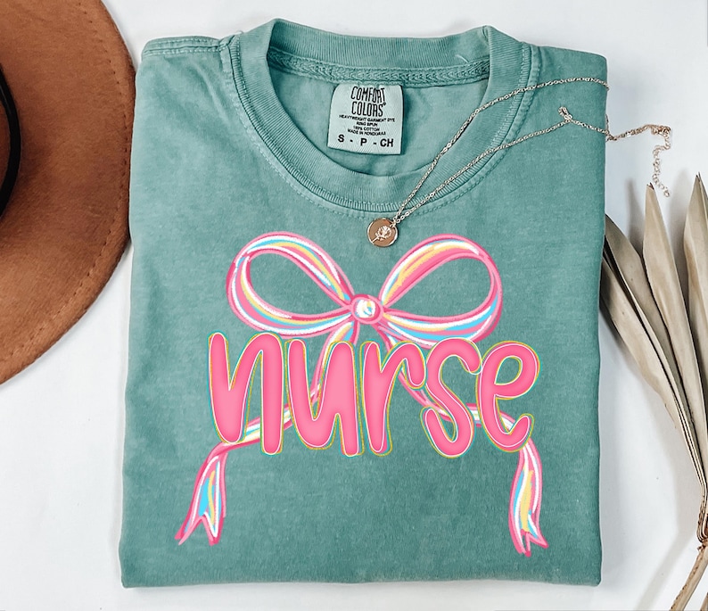 Pode incluir: Uma camiseta Comfort Colors verde-azulado com um design de la&ccedil;o rosa e arco-&iacute;ris e a palavra "nurse" em rosa. Um colar de ouro com um pequeno pingente est&aacute; drapeado sobre a camisa.