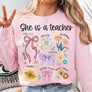 Op de afbeelding: Roze sweatshirt met de tekst "She is a teacher" in het zwart. Het ontwerp bevat bloemen- en lintafbeeldingen, samen met inspirerende woorden als "Chosen", "Beautiful" en "Victorious".
