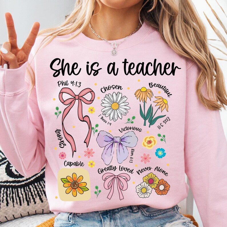Op de afbeelding: Roze sweatshirt met de tekst "She is a teacher" in het zwart. Het ontwerp bevat bloemen- en lintafbeeldingen, samen met inspirerende woorden als "Chosen", "Beautiful" en "Victorious".