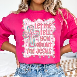 May include: A bright pink sweatshirt with the text "Let me tell you about my Jesus" in a decorative frame. The graphic includes a cross with a bow and pastel colors.