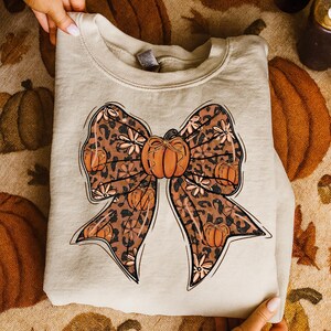 May include: A beige sweatshirt featuring a large bow design with a pumpkin in the center. The bow has a leopard print pattern with orange pumpkins and white floral accents. The sweatshirt is held by hands.