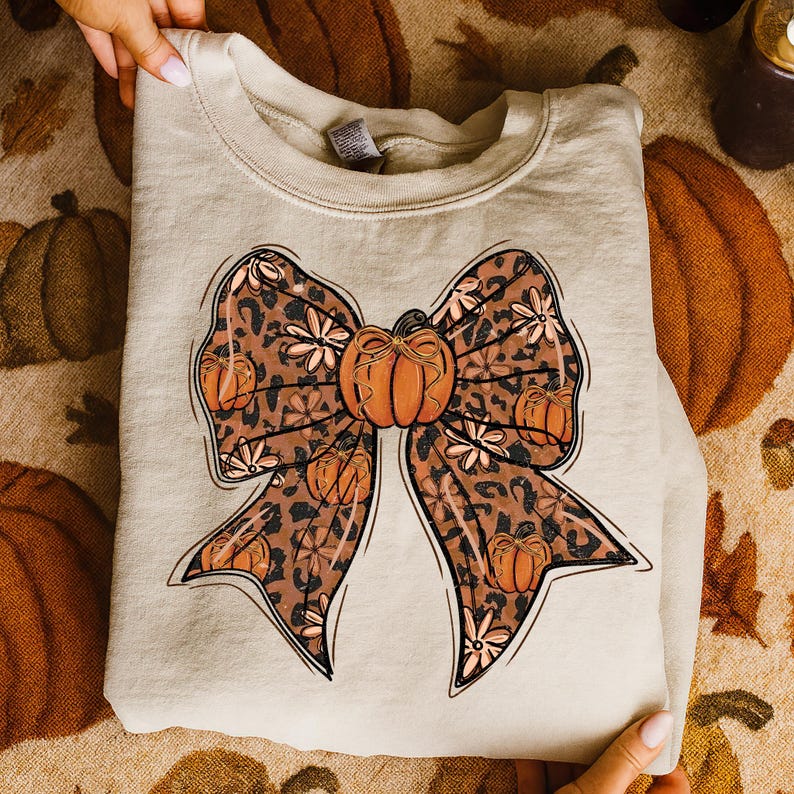 May include: A beige sweatshirt featuring a large bow design with a pumpkin in the center. The bow has a leopard print pattern with orange pumpkins and white floral accents. The sweatshirt is held by hands.