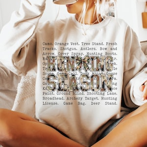 May include: A beige sweatshirt with the words "HUNTING SEASON" in a camouflage pattern. The sweatshirt also lists hunting-related terms such as "Shotgun," "Antlers," and "Hunting Boots."