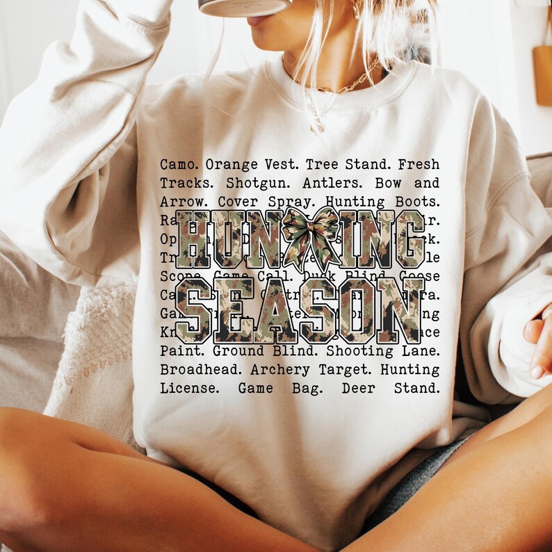 May include: A beige sweatshirt with the words "HUNTING SEASON" in a camouflage pattern. The sweatshirt also lists hunting-related terms such as "Shotgun," "Antlers," and "Hunting Boots."