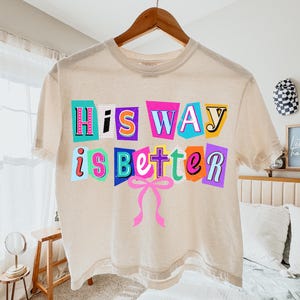 May include: A beige t-shirt with the phrase "HIS WAY is better" in colorful, cut-out style letters. A pink bow is below the text. The shirt is hanging on a wooden hanger.