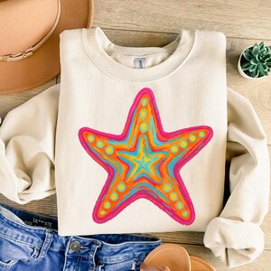 May include: Cream-colored sweatshirt featuring a vibrant starfish design. The starfish is outlined in pink, with orange, yellow, and blue details. A casual, comfortable top for beach lovers.