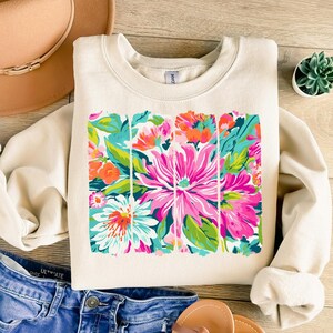 May include: A cream-colored sweatshirt with a floral print design. The print features a variety of pink, green, blue, and orange flowers. The flowers are arranged in a rectangular shape with a distressed, painted look.