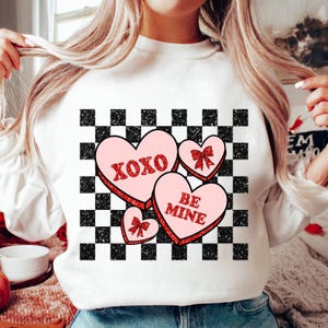 May include: White sweatshirt with a black and white checkered pattern and pink hearts with red glitter text that says "XOXO" and "BE MINE".