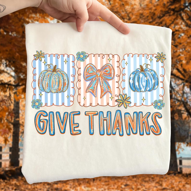 May include: Cream-colored t-shirt featuring a fall-themed design. The design includes three framed sections with pumpkins and a bow, set against striped backgrounds. The words "GIVE THANKS" are printed in a colorful, outlined font.