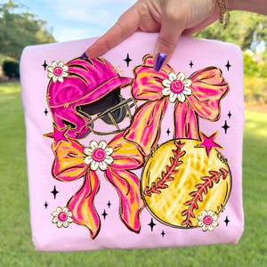 May include: Pink sweatshirt featuring a softball helmet, softball, and bows in pink and yellow. The design includes star and flower accents. The softball is yellow with red stitching. The bows have a gold outline.