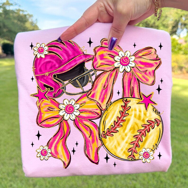 May include: Pink sweatshirt featuring a softball helmet, softball, and bows in pink and yellow. The design includes star and flower accents. The softball is yellow with red stitching. The bows have a gold outline.