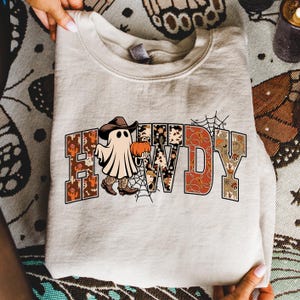 Puede incluir: Una sudadera beige con la palabra "HOWDY" en una fuente decorativa. Los dise&ntilde;os de las letras presentan una variedad de patrones, incluyendo un fantasma con sombrero y botas de vaquero, una calabaza y una telara&ntilde;a. La sudadera est&aacute; doblada.