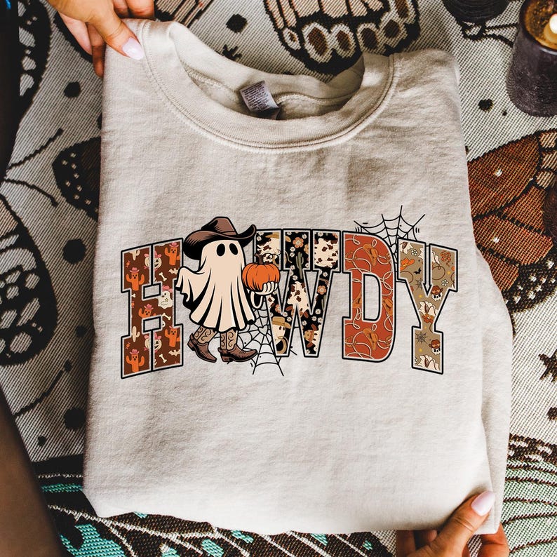 Puede incluir: Una sudadera beige con la palabra "HOWDY" en una fuente decorativa. Los dise&ntilde;os de las letras presentan una variedad de patrones, incluyendo un fantasma con sombrero y botas de vaquero, una calabaza y una telara&ntilde;a. La sudadera est&aacute; doblada.