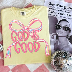 May include: A light yellow t-shirt with the text "GOD IS GOOD" in pink, outlined in a rainbow of colors. The design includes a bow. A newspaper and a disco ball are in the background.