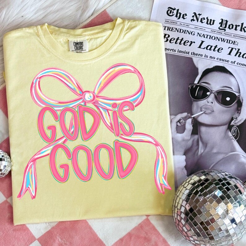 May include: A light yellow t-shirt with the text "GOD IS GOOD" in pink, outlined in a rainbow of colors. The design includes a bow. A newspaper and a disco ball are in the background.
