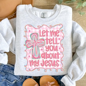 May include: A light gray sweatshirt with the text "Let me tell you about my Jesus" in pink and blue. The design includes a cross with a pink bow and a pink gingham background.