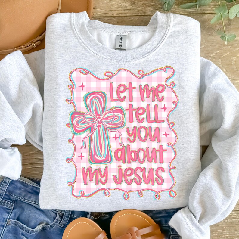 May include: A light gray sweatshirt with the text "Let me tell you about my Jesus" in pink and blue. The design includes a cross with a pink bow and a pink gingham background.