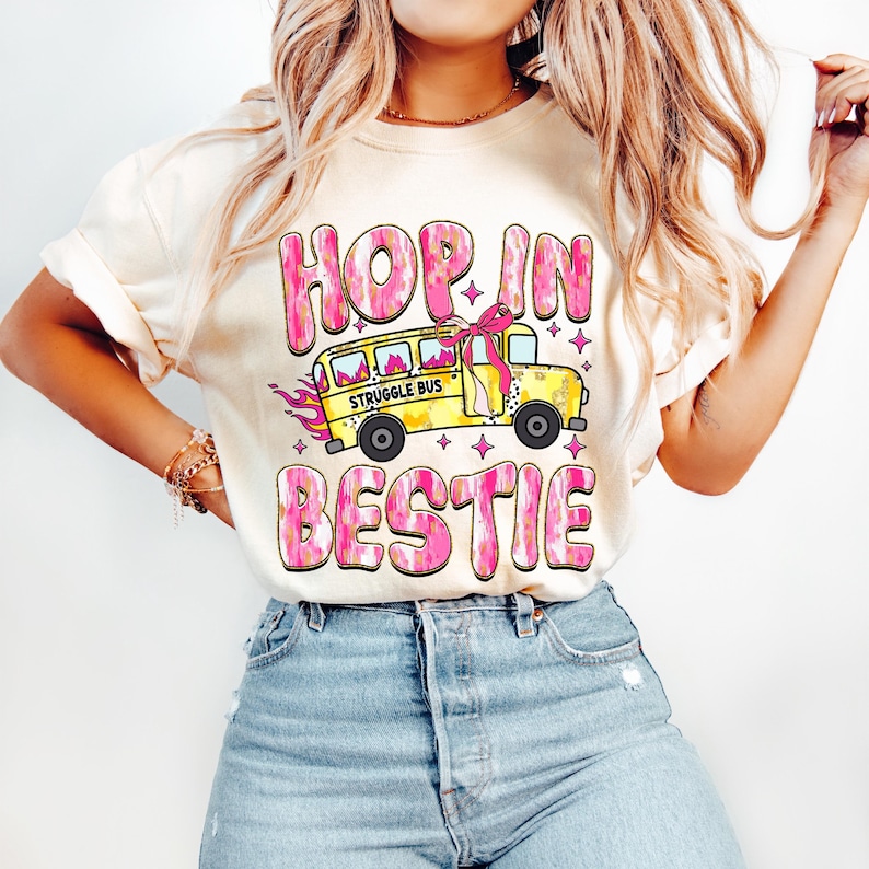 May include: A white t-shirt with a pink and yellow graphic design. The graphic features a yellow school bus with the words "Struggle Bus" on the side. The bus has a pink bow on top and is surrounded by pink and yellow stars. The text "Hop in bestie" is written in pink and yellow letters above the bus.