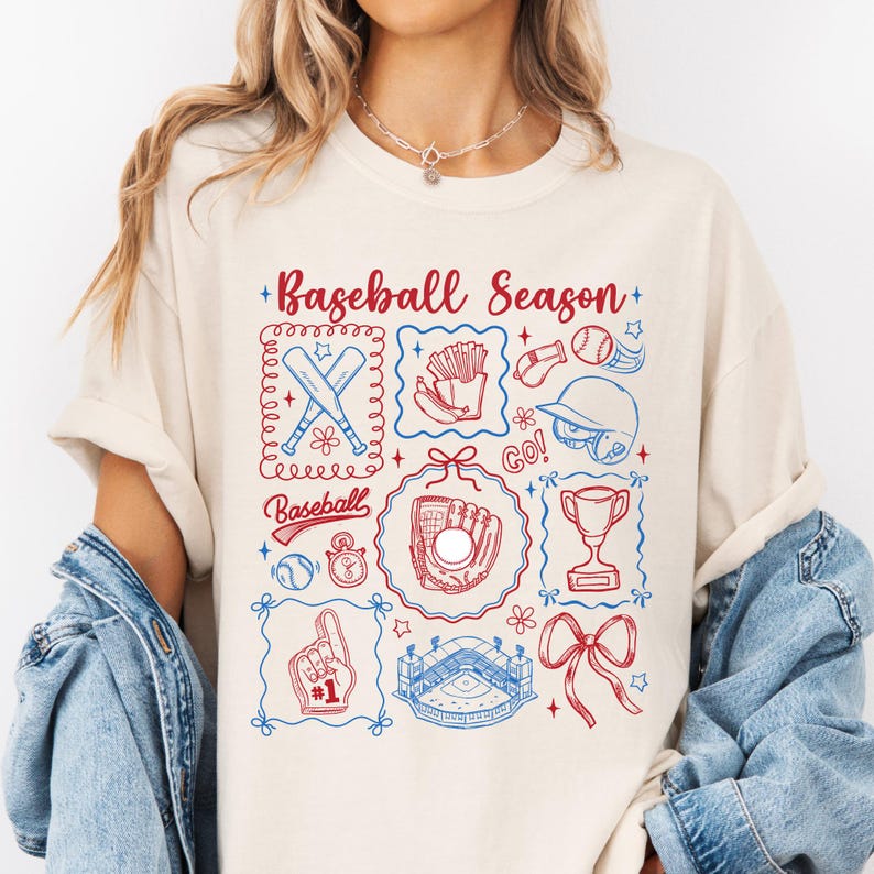 May include: Cream-colored sweatshirt with a baseball-themed graphic. The design includes baseball bats, a glove, a helmet, a trophy, and the words "Baseball Season" in red and blue. The graphic is surrounded by decorative frames.