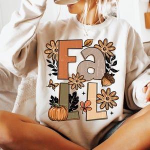 May include: Cream-colored sweatshirt with the word "FALL" in a fall-themed design. The letters are in shades of brown, tan, and green, with floral and leaf accents, a pumpkin, butterfly, and acorn.