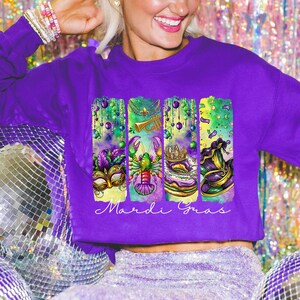 May include: Purple sweatshirt with a graphic design of Mardi Gras themed elements, including a mask, a crawfish, a king cake, and a jester's hat. The text "Mardi Gras" is printed below the graphic.