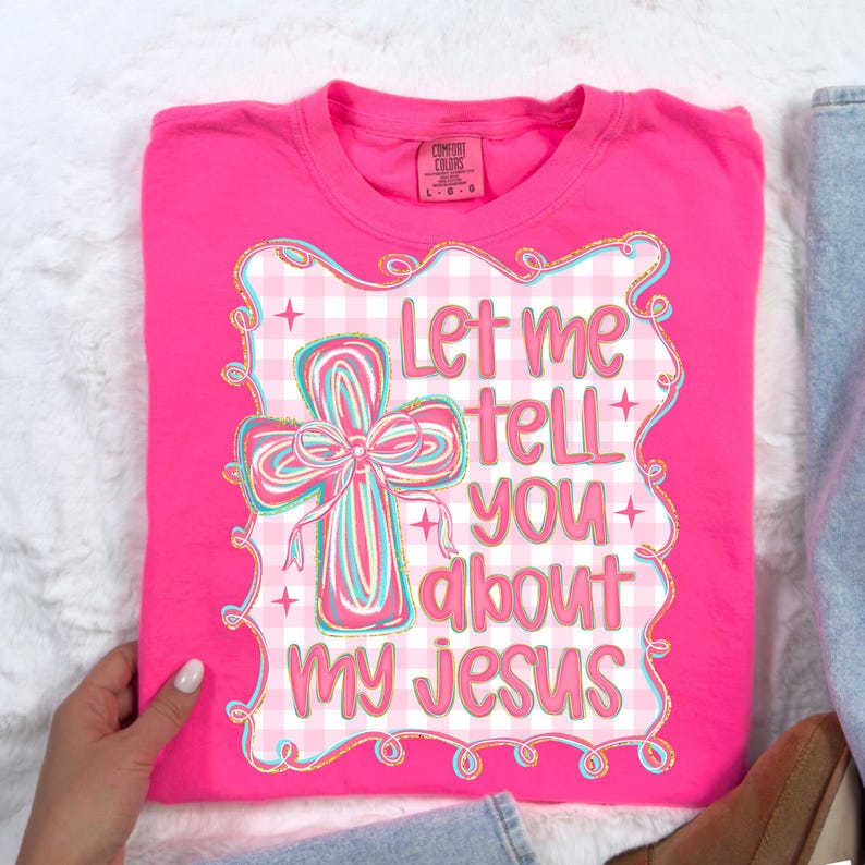 May include: A hot pink t-shirt with the text "Let me tell you about my Jesus" in a decorative font. The design includes a cross with a bow and a gingham pattern, outlined in gold.
