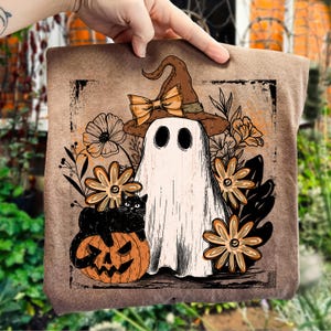 May include: A brown Halloween-themed pillow featuring a ghost wearing a witch hat with a bow, a black cat, and a jack-o'-lantern. The design includes orange and cream-colored flowers and foliage, all set against a textured background.