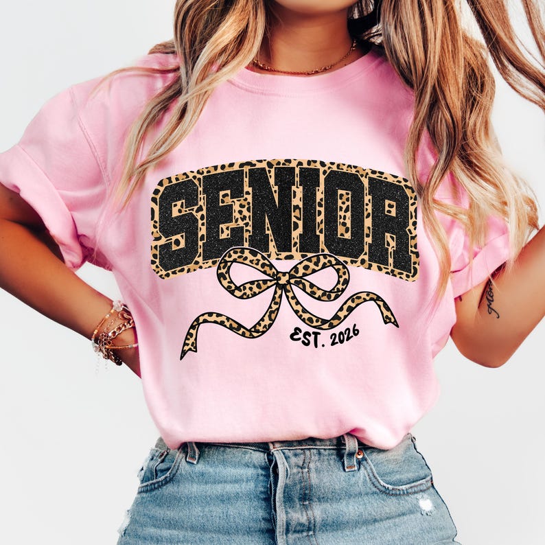 May include: A light pink t-shirt with the word "SENIOR" in large, black letters with a leopard print fill. Below the word is a leopard print bow with the text "EST. 2026".