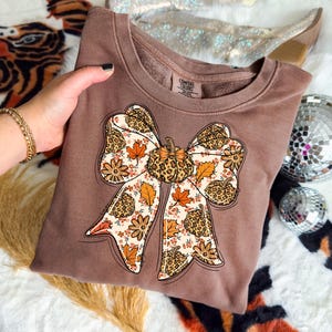May include: Brown sweatshirt with a large bow design. The bow features a pumpkin in the center and is decorated with fall leaves, flowers, and a leopard print pattern. The sweatshirt has a crew neck and long sleeves.