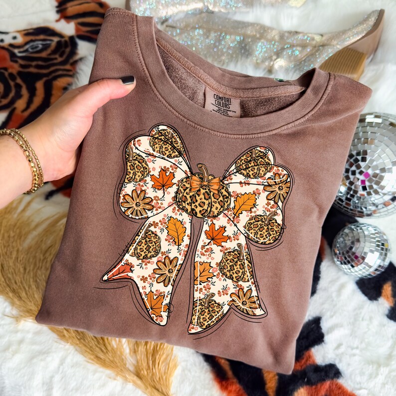 May include: Brown sweatshirt with a large bow design. The bow features a pumpkin in the center and is decorated with fall leaves, flowers, and a leopard print pattern. The sweatshirt has a crew neck and long sleeves.