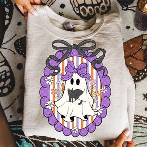 May include: A beige sweatshirt featuring a Halloween-themed graphic. The graphic includes a ghost with a black cat, surrounded by flowers, inside a purple oval frame with a black bow. The background has orange and white stripes.