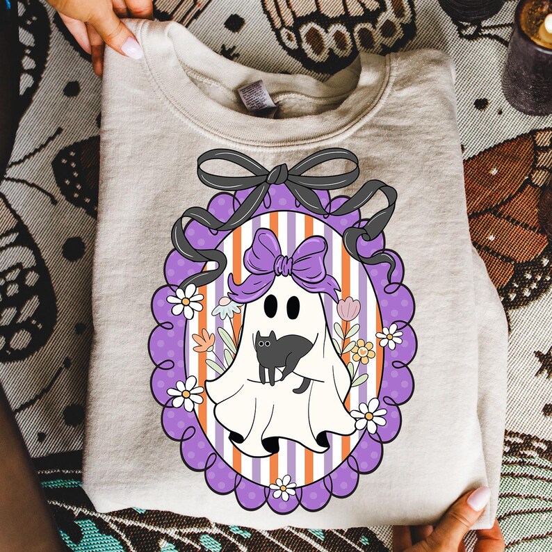 May include: A beige sweatshirt featuring a Halloween-themed graphic. The graphic includes a ghost with a black cat, surrounded by flowers, inside a purple oval frame with a black bow. The background has orange and white stripes.