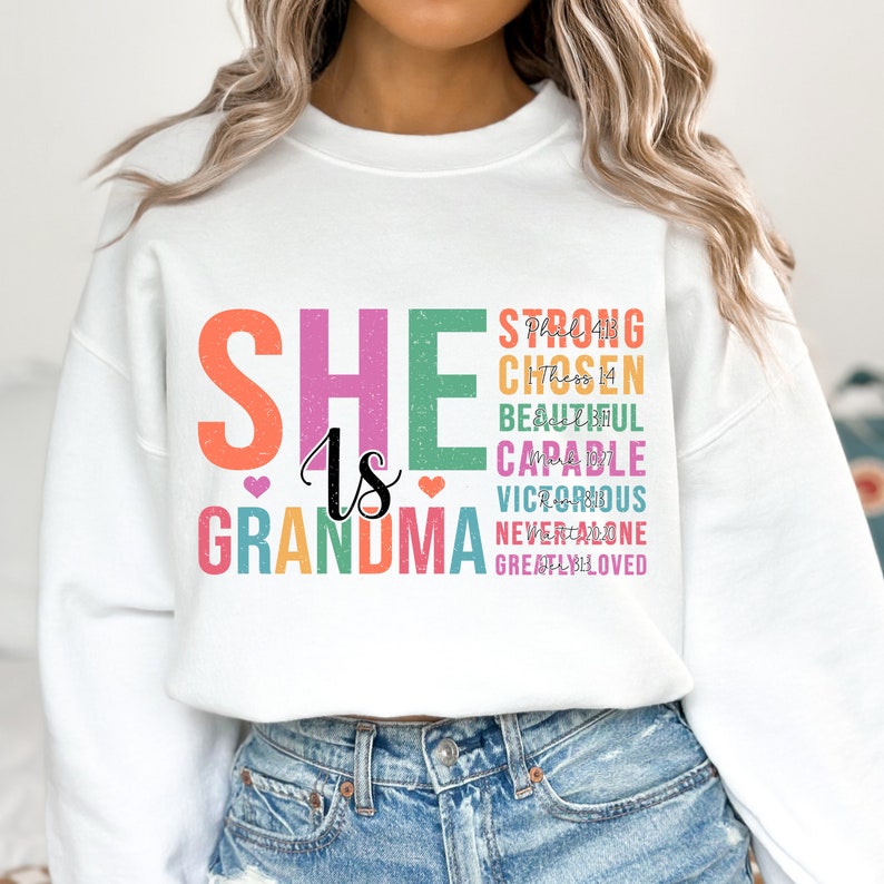 May include: White sweatshirt with the text "SHE is GRANDMA" in colorful letters. The text is stacked vertically with the words "STRONG, CHOSEN, BEAUTIFUL, CAPABLE, VICTORIOUS, NEVER ALONE, GREATLY LOVED" stacked vertically to the right of the main text.