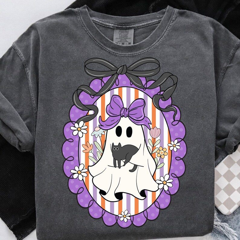 May include: Dark gray sweatshirt featuring a Halloween-themed design. The graphic includes a white ghost with a purple bow, a black cat, and floral accents within a purple and orange striped oval frame, topped with a black bow.