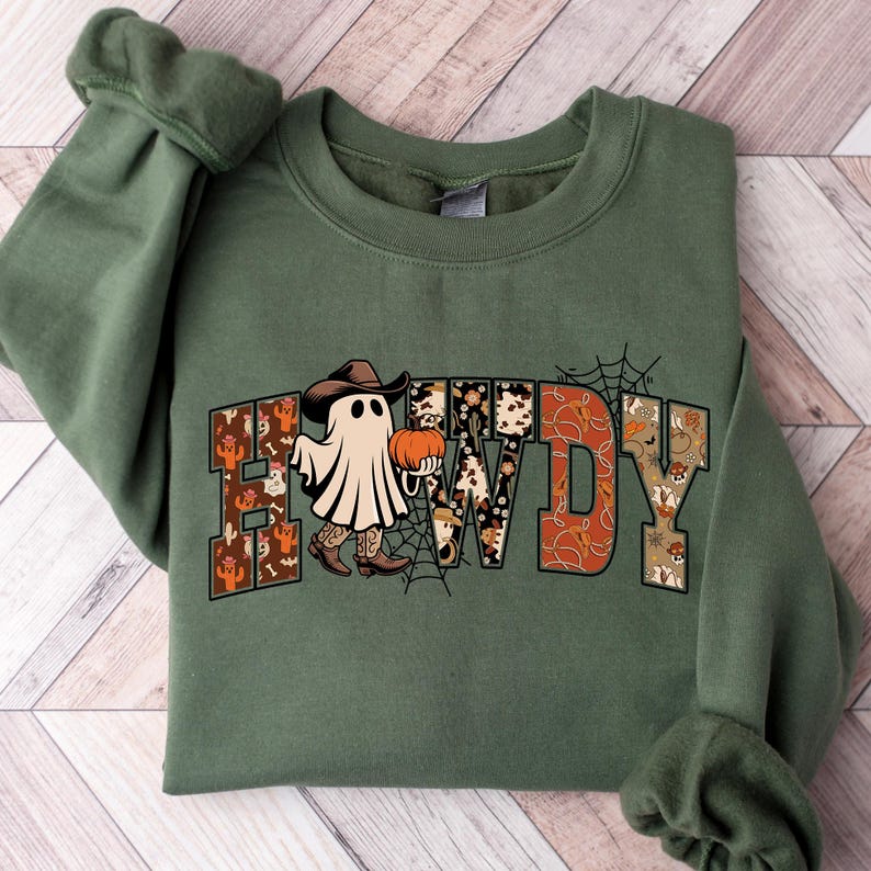 Puede incluir: Sudadera verde con la palabra "HOWDY" en una fuente de estilo occidental. Las letras est&aacute;n decoradas con patrones de calabazas, cactus y piel de vaca. Un fantasma con sombrero y botas de vaquero sostiene una calabaza. Una telara&ntilde;a est&aacute; en el fondo.