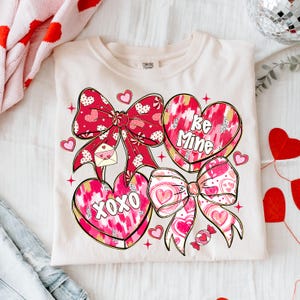 Puede incluir: Camiseta color crema con un dise&ntilde;o de San Valent&iacute;n. El gr&aacute;fico incluye lazos rojos, caramelos en forma de coraz&oacute;n con las frases "Be Mine" y "XOXO", y peque&ntilde;os corazones. El dise&ntilde;o tiene un tema juguet&oacute;n y rom&aacute;ntico.
