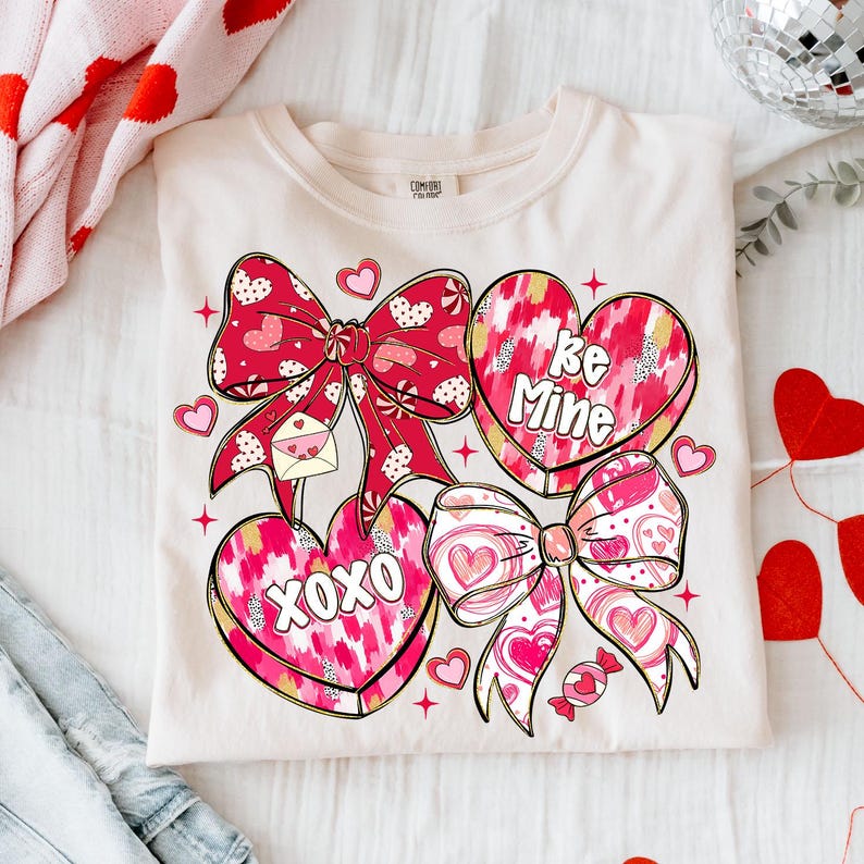 Puede incluir: Camiseta color crema con un dise&ntilde;o de San Valent&iacute;n. El gr&aacute;fico incluye lazos rojos, caramelos en forma de coraz&oacute;n con las frases "Be Mine" y "XOXO", y peque&ntilde;os corazones. El dise&ntilde;o tiene un tema juguet&oacute;n y rom&aacute;ntico.