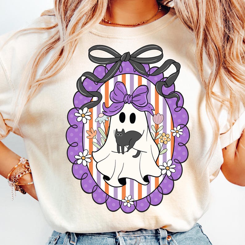 May include: Cream-colored t-shirt featuring a Halloween-themed graphic. The design includes a white ghost with a black cat, a purple bow, and a decorative frame with flowers. The background has vertical stripes in orange, white, and purple.