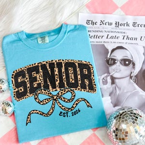 May include: A light blue t-shirt with the word "SENIOR" in a black glitter and leopard print design. A leopard print bow is below the word "SENIOR" with the text "EST. 2026". A newspaper clipping and disco ball are in the background.
