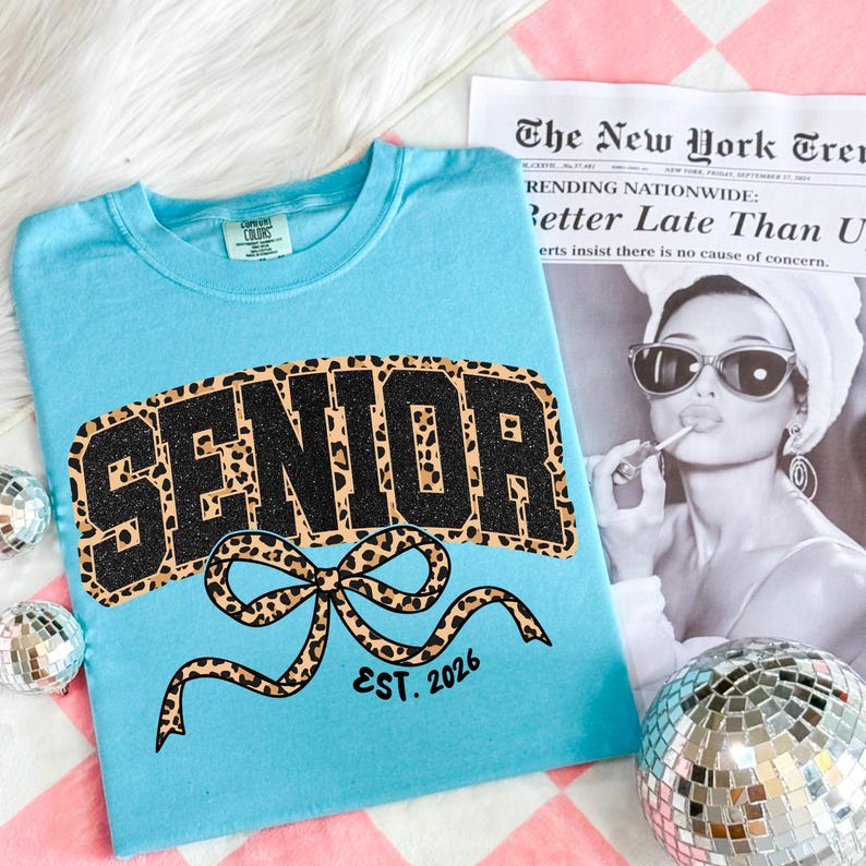 May include: A light blue t-shirt with the word "SENIOR" in a black glitter and leopard print design. A leopard print bow is below the word "SENIOR" with the text "EST. 2026". A newspaper clipping and disco ball are in the background.
