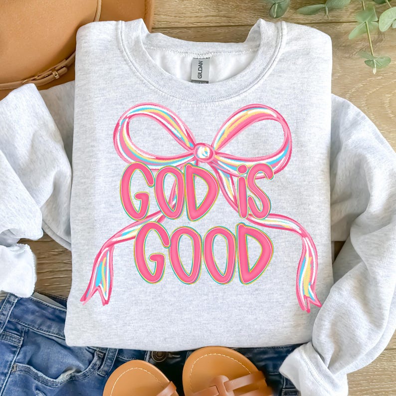 May include: A light gray sweatshirt with the words "GOD IS GOOD" in pink, outlined in a darker pink. The text is designed to look like a ribbon. The ribbon has a rainbow design. The sweatshirt is folded on a surface with other items.