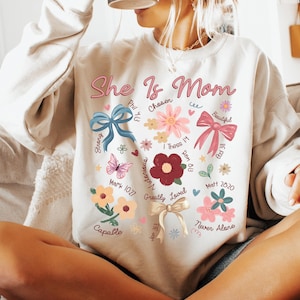 K&ouml;nnte beinhalten: Ein wei&szlig;er Pullover mit einem herzf&ouml;rmigen Design, das die Worte "She Is Mom" zeigt, umgeben von Blumen, Schmetterlingen und Schleifen. Das Design enth&auml;lt auch Bibelverse und W&ouml;rter wie "Chosen", "Strong", "Victorious", "Greatly Loved", "Capable" und "Never Alone".