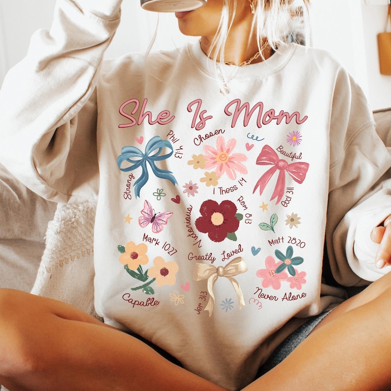 K&ouml;nnte beinhalten: Ein wei&szlig;er Pullover mit einem herzf&ouml;rmigen Design, das die Worte "She Is Mom" zeigt, umgeben von Blumen, Schmetterlingen und Schleifen. Das Design enth&auml;lt auch Bibelverse und W&ouml;rter wie "Chosen", "Strong", "Victorious", "Greatly Loved", "Capable" und "Never Alone".