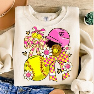 May include: Cream-colored sweatshirt featuring a softball-themed graphic. The design includes a yellow softball, pink helmet, glove, bows, and flowers. The bows have a pink and yellow checkered pattern, and the flowers have pink centers.