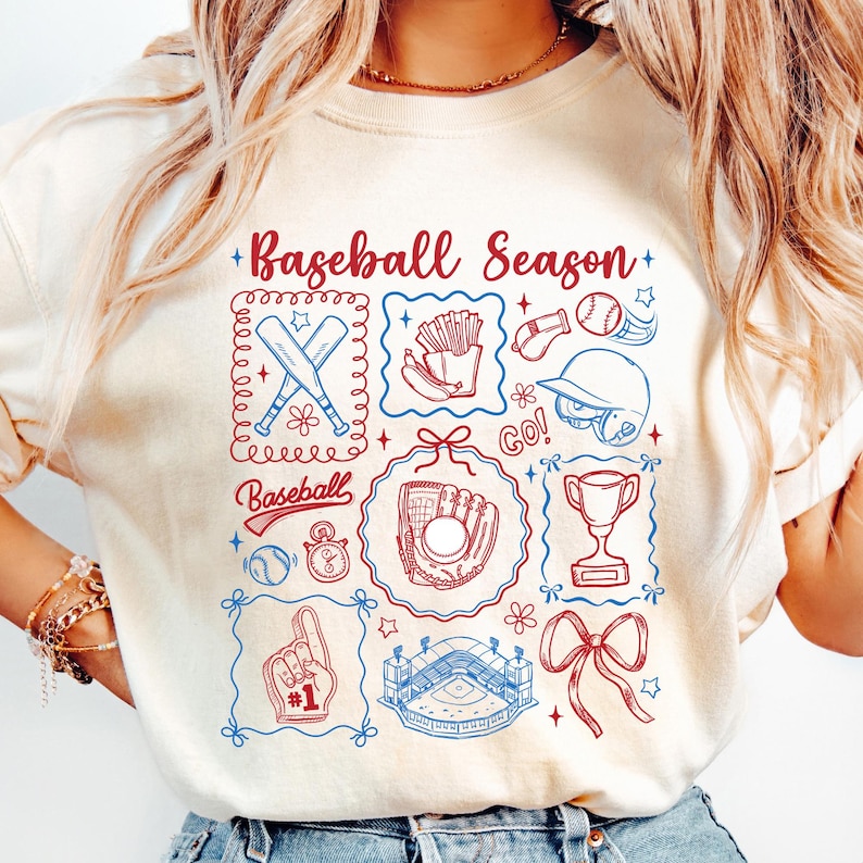 May include: Cream-colored t-shirt with a baseball-themed graphic. The design includes baseball bats, a glove, a stadium, and the text "Baseball Season." The graphic is in red and blue, with a decorative border.