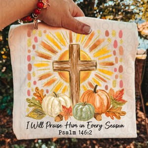 May include: A cream-colored t-shirt featuring a graphic of a wooden cross centered before a yellow sunburst. Below the cross are white and green pumpkins, autumn leaves, and acorns. The text reads: I Will Praise Him in Every Season Psalm 146:2.