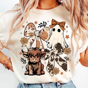 Op de afbeelding: Cr&egrave;mekleurig grafisch T-shirt met een Schotse hooglander, een spook en strikken met bloemen- en koeienhuidpatronen. Het ontwerp bevat oranje sterren en bloemen, wat een grillige, herfstachtige esthetiek cre&euml;ert.