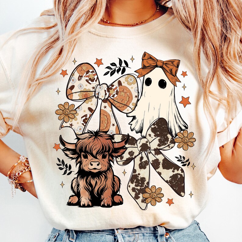 Op de afbeelding: Cr&egrave;mekleurig grafisch T-shirt met een Schotse hooglander, een spook en strikken met bloemen- en koeienhuidpatronen. Het ontwerp bevat oranje sterren en bloemen, wat een grillige, herfstachtige esthetiek cre&euml;ert.