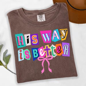May include: Brown Comfort Colors t-shirt with the phrase "His Way is Better" in colorful, cut-out style letters. A pink bow is below the text. The shirt is folded on a white surface.
