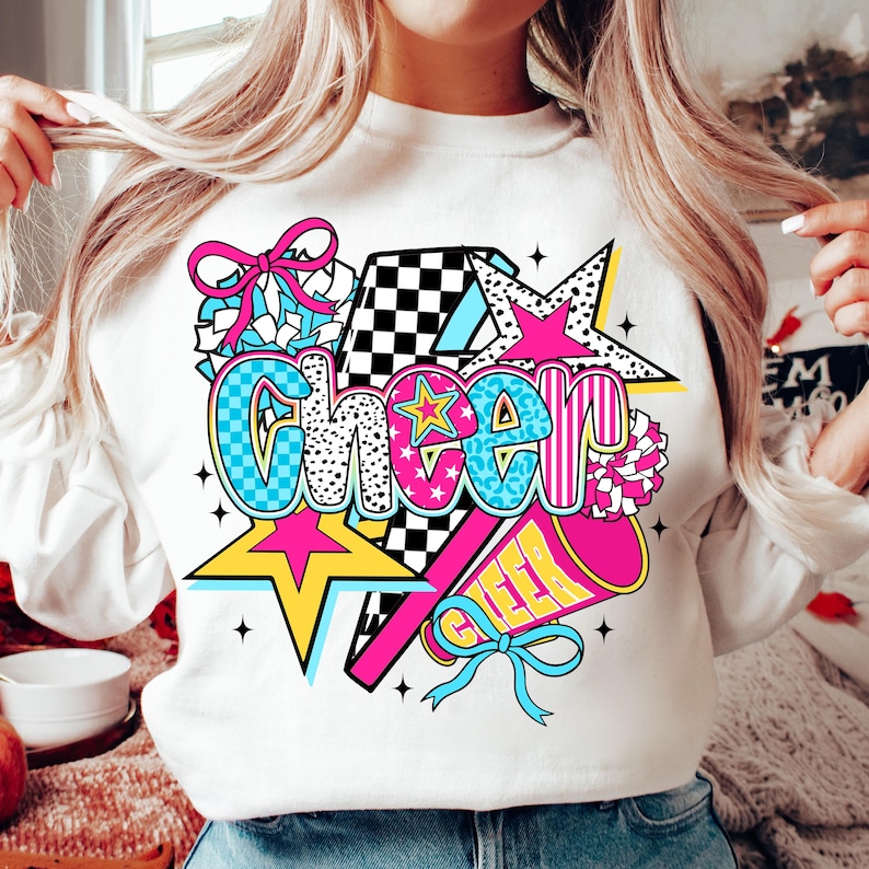 Op de afbeelding: Witte sweatshirt met een kleurrijke "Cheer" afbeelding. Het ontwerp bevat een ster, pompon, megafoon en strik. Het woord "Cheer" is in verschillende lettertypen en patronen, tegen een geblokte achtergrond.